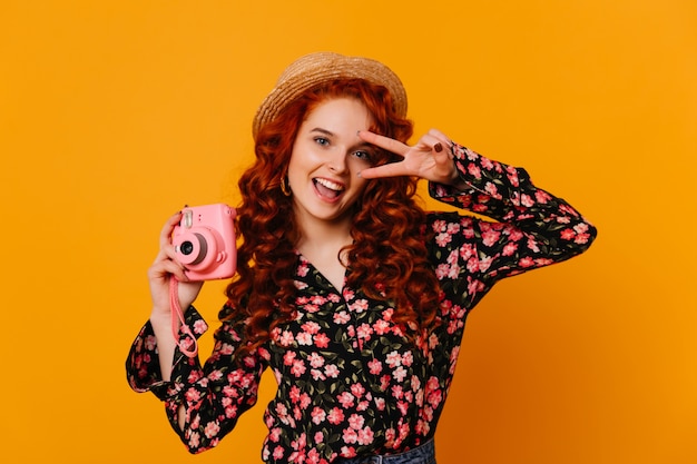 Blauäugige süße Frau mit rotem Haar lächelt und prickelt Friedenszeichen und hält rosa Kamera auf isoliertem Raum.