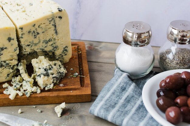 Blauschimmelkäse mit Oliven; Salz- und Pfefferstreuer mit Serviette auf Schreibtisch aus Holz