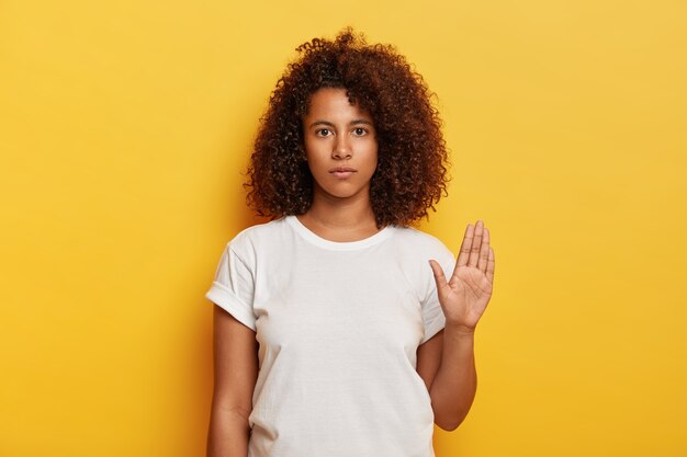 Bleib mir fern. Ernstes attraktives Mädchen mit lockigem Haar, hält Handfläche in Stopp-Geste, sieht selbstbewusst aus, trägt weißes T-Shirt, isoliert auf gelber Wand, sagt, es ist genug. Körpersprache