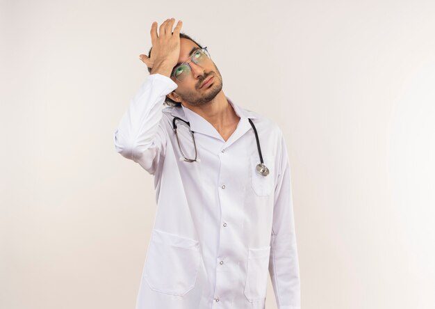 Blick auf den denkenden jungen männlichen Arzt mit optischer Brille in weißem Gewand mit Stethoskop