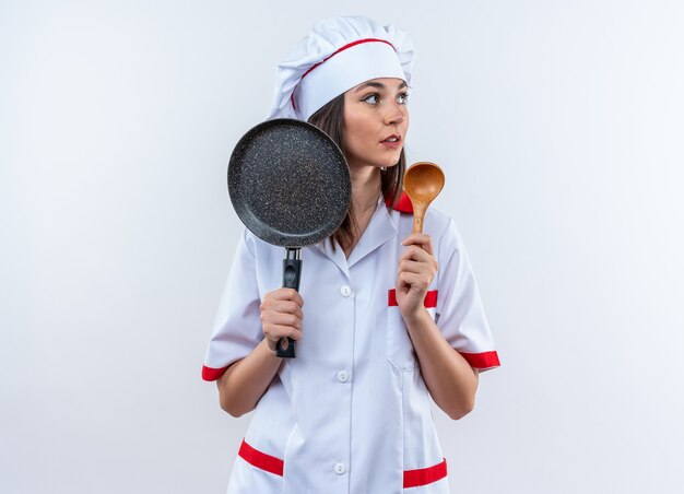 Blick auf die Seite junge Köchin in Kochuniform mit Bratpfanne mit Löffel isoliert auf weißer Wand