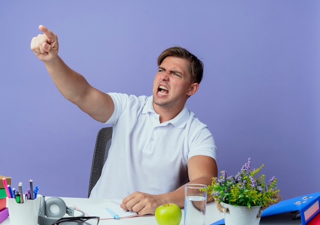 Blick auf die Seite wütend junger hübscher männlicher Student, der am Schreibtisch mit den Schulwerkzeugpunkten an der Seite sitzt