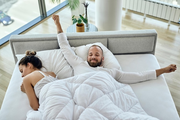 Blick aus der vogelperspektive auf einen glücklichen mann, der sich morgens streckt, während er mit seiner freundin im bett liegt