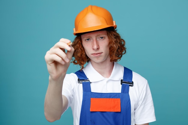 Blick in die Kamera junger Baumeister in Uniform, der einen Markierungsstift in die Kamera hält, isoliert auf blauem Hintergrund