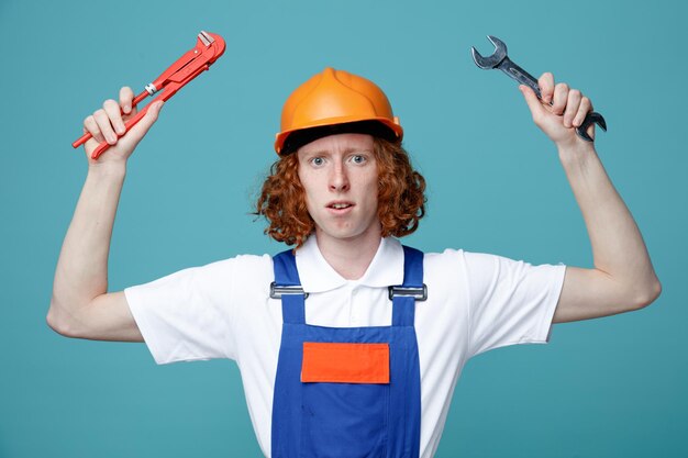 Blick in die Kamera junger Baumeister in Uniform mit Schraubenschlüsseln am Hals isoliert auf blauem Hintergrund