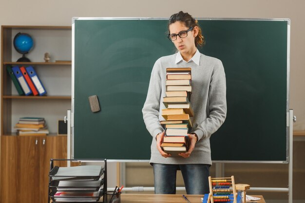 Kostenloses Foto blick in die kamera junger männlicher lehrer, der bücher hält, die vor der tafel im klassenzimmer stehen