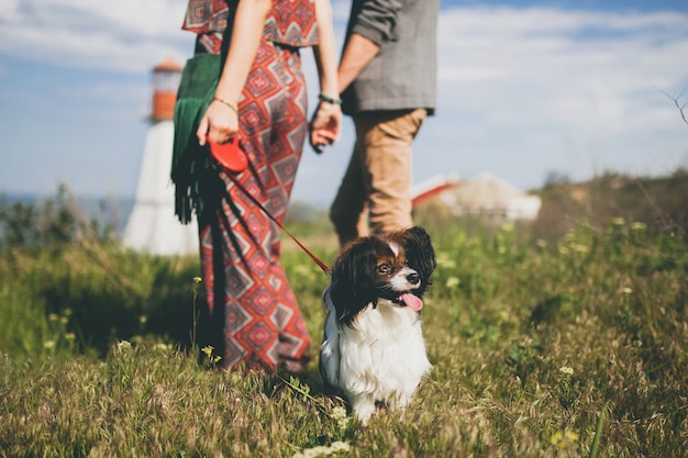 Blick von hinten auf junges stilvolles Hipster-Paar in der Liebe, das mit Hund in der Landschaft geht