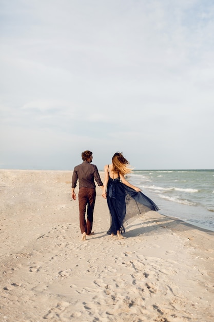Blick von hinten. Elegantes verliebtes Paar, das entlang des Strandes geht. Romantische Momente. Weißer Sand und Meereswellen. Tropischer Urlaub. Vollständige Höhe.
