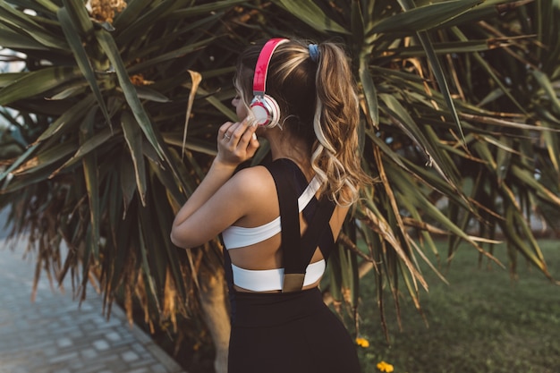 Blick von hinten freudige erstaunliche Frau in Sportbekleidung, mit langen lockigen Haaren, die Musik über Kopfhörer in tropischer Stadt hören. Sonniger Morgen, Palmen, wahre Emotionen, gesunder Lebensstil, Training.