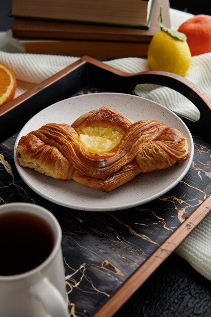 Blick von oben auf das Frühstücksset mit Croissant auf Teller und Marmelade in Schüssel und Tasse Tee auf Tablett mit Orangen- und Zitronenscheiben mit offenem Buch und kleiner Blume auf Buch auf Stoff auf schwarzem Hintergrund