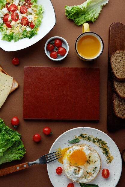 Blick von oben auf das Frühstücksset mit Spiegelei mit Tomaten und Dill und Gemüsesalat mit Tomaten-Ei-Salat mit Gabel-Orangensaft-Brotscheiben und Schneidebrett in der Mitte