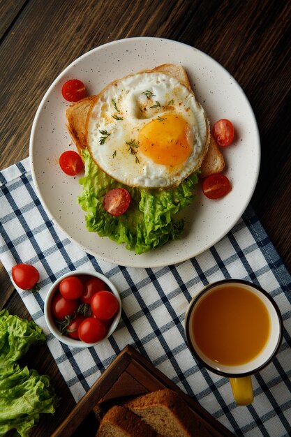 Blick von oben auf das Frühstücksset mit Spiegelei-Salat-Tomaten auf getrockneter Brotscheibe im Teller mit Orangensaft auf kariertem Stoff und Schachtel Brotscheiben auf Holzhintergrund