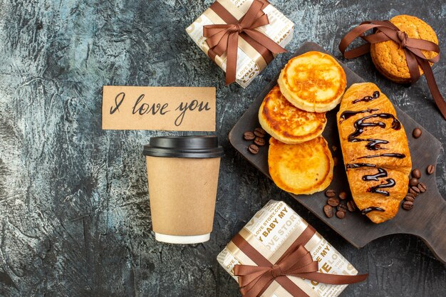 Blick von oben auf das Schneidebrett mit leckerem Frühstück mit Pfannkuchen Croisasant gestapelten Keksen schöne Geschenkboxen Kaffee auf dunkler Oberfläche on