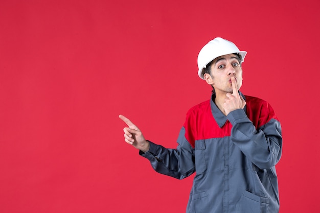 Blick von oben auf den jungen Architekten in Uniform mit Schutzhelm, der auf die rechte Seite auf isolierte rote Wand zeigt