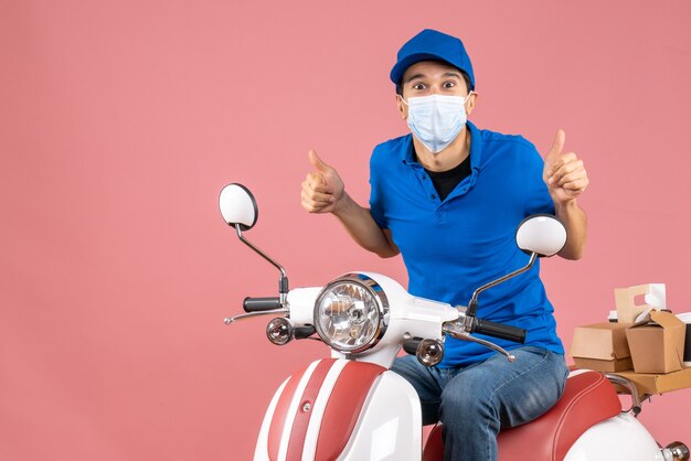 Blick von oben auf den selbstbewussten Lieferer in medizinischer Maske mit Hut, der auf dem Roller sitzt und eine gute Geste auf pastellfarbenem Pfirsichhintergrund macht