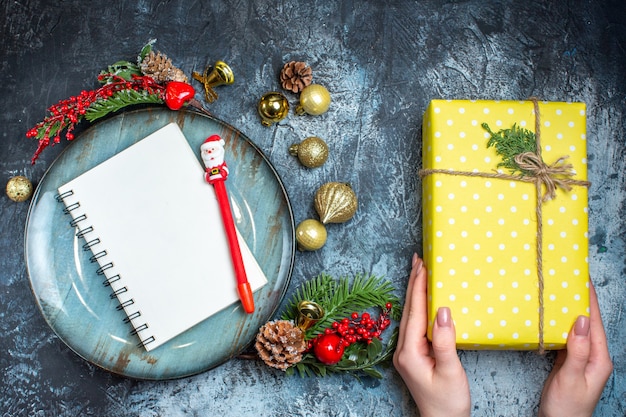 Blick von oben auf die Hand, die eine Geschenkbox und ein Notizbuch mit blauem Teller mit dekorativem Zubehör Nadelbaumkegel auf dunklem Hintergrund hält