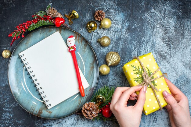 Blick von oben auf die Hand, die eine Geschenkbox und ein Notizbuch mit dekorativem Zubehör des Stiftes blaue Platte öffnet Nadelbaumkegel auf dunklem Hintergrund