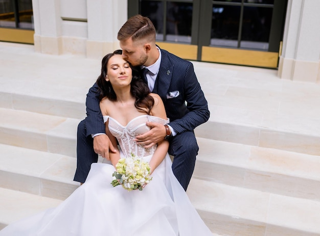 Kostenloses Foto blick von oben auf einen eleganten mann im stilvollen anzug, der auf einer treppe vor dem hintergrund eines gebäudes sitzt und die braut umarmt und küsst, die einen blumenstrauß in der hand hält und sich glücklich fühlt