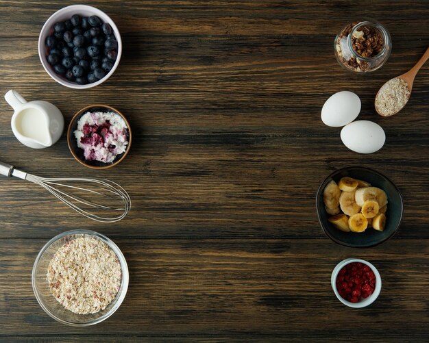 Blick von oben auf Frühstückszutaten wie Banane, Hafer, Ei, Walnuss, Milch, Hüttenkäse, rote Johannisbeere, Schwarzdorn mit Schneebesen auf Holzhintergrund mit Kopierraum