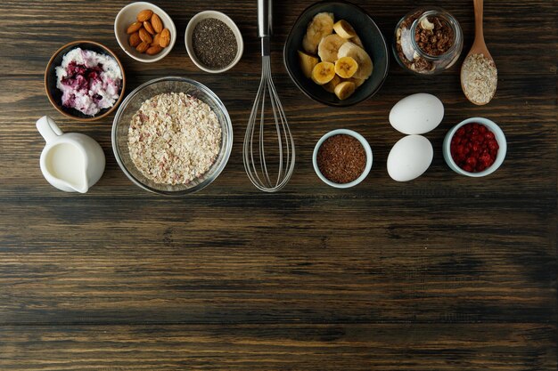 Blick von oben auf Frühstückszutaten wie Banane, Hafer, Ei, Walnuss, Sesam, Schwarzdorn, Milch, Hüttenkäse, Mandel, rote Johannisbeere mit Schneebesen auf Holzhintergrund mit Kopierraum