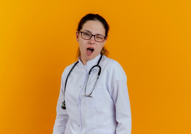 blinkende junge Ärztin, die medizinische Robe und Stethoskop mit isolierter Brille trägt