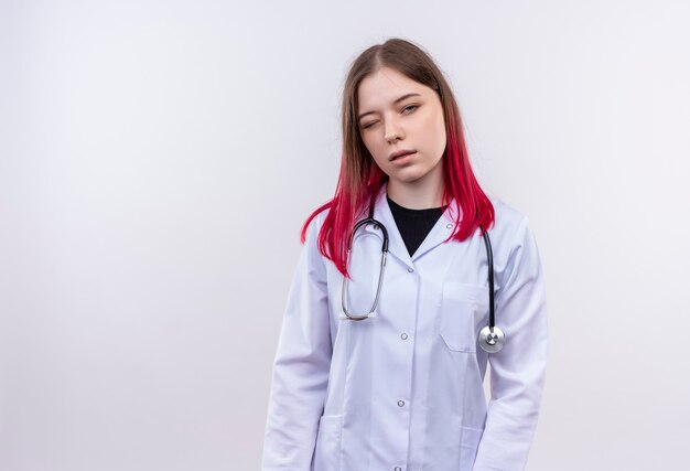 Blinkende junge Ärztin, die medizinisches Gewand des Stethoskops auf isolierter weißer Wand mit Kopienraum trägt