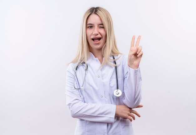Blinkendes freudiges freudiges junges blondes Mädchen des Arztes, das Stethoskop und medizinisches Kleid in Zahnspange trägt, die Friedensgeste auf isolierter weißer Wand zeigt