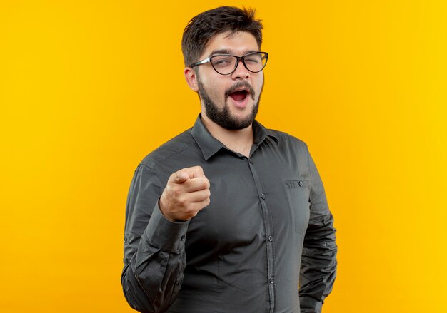 Blinzelte freudiger junger Geschäftsmann, der Brille trägt, die Sie Geste lokalisiert auf gelbem Hintergrund zeigt