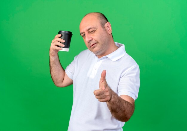 Blinzelte lässiger reifer Mann, der Tasse Kaffee hält und Ihnen gestikulierte Geste auf grüner Wand zeigt