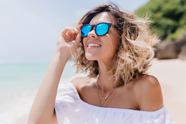 Blithesome blonde Frau in der Sonnenbrille, die Himmel betrachtet. Außenporträt der bezaubernden kaukasischen Frau, die am Sandstrand kühlt.