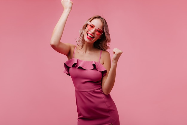 Blithesome europäische junge Frau in der Sonnenbrille, die auf rosiger Wand tanzt