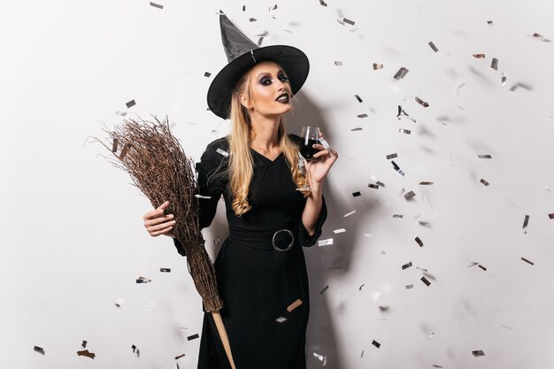 Blithesome junge Hexe, die Wein trinkt. Sorglose Frau in Halloween-Kleidung, die auf Partei aufwirft.