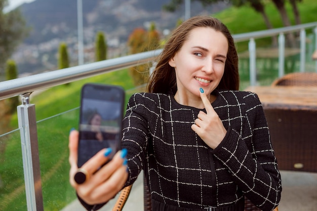 Blogger-Mädchen macht Selfie mit der Rückkamera des Smartphones, indem sie den Zeigefinger auf dem Kinn auf dem Hintergrund der Naturansicht hält