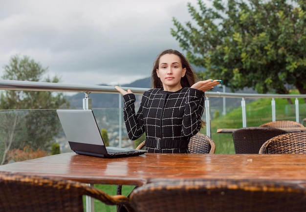 Blogger-Mädchen mit Laptop schaut in die Kamera, indem sie auf dem Hintergrund der Naturansicht sitzt