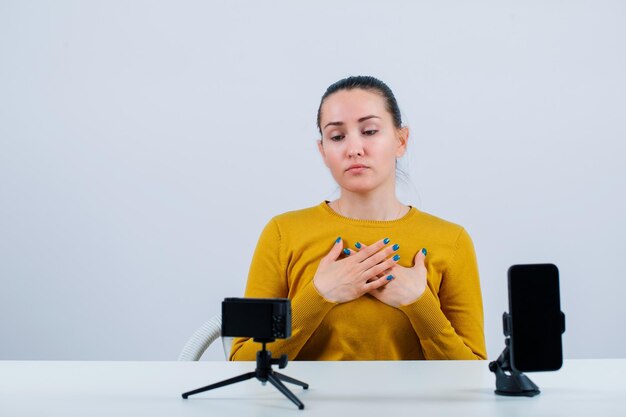 Blogger-Mädchen posiert, indem sie die Hände auf der Brust vor ihrer Mini-Kamera auf weißem Hintergrund hält
