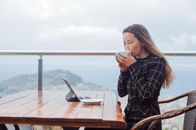 Blogger-Mädchen schaut auf den Laptop-Bildschirm, indem sie eine Tasse heißen Kaffee auf dem Hintergrund der Stadtansicht trinkt