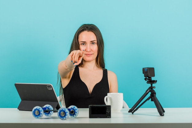 Blogger-Mädchen schaut in die Kamera, indem sie den Kamerafokus mit dem Zeigefinger auf blauen Hintergrund zeigt