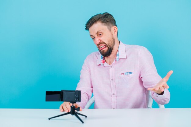 Blogger-Mann posiert vor seiner Minikamera, indem er lustige Mimikry auf blauem Hintergrund zeigt