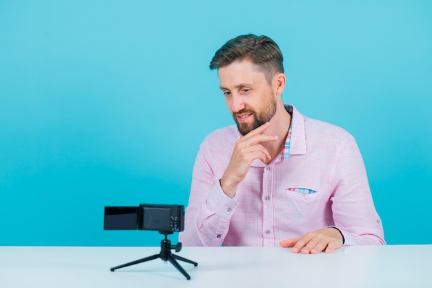 Blogger-Mann schaut auf seinen Mini-Kamerabildschirm, indem er die Hand am Kinn auf blauem Hintergrund hält