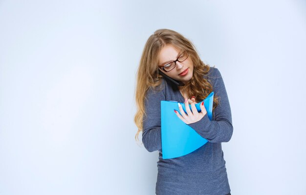 Blonde Assistentin, die mit dem Telefon spricht, während sie ihren blauen Ordner überprüft und Korrekturen vornimmt.