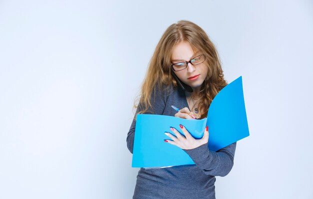 Blonde Assistentin, die mit dem Telefon spricht, während sie ihren blauen Ordner überprüft und Korrekturen vornimmt.