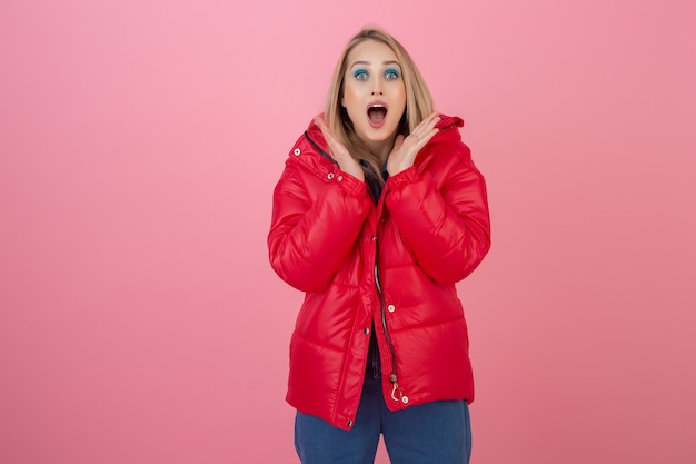 Blonde attraktive aktive Frau, die auf rosa Wand in der bunten Winter-Daunenjacke der roten Farbe aufwirft, Spaß hat, warmen Mantelmodetrend, überraschten schockierten Gesichtsausdruck