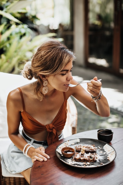 Blonde charmante Frau in braunem BH und Jeansshorts isst Waffel mit Sahne und Schokoladensauce und genießt ihren Geschmack