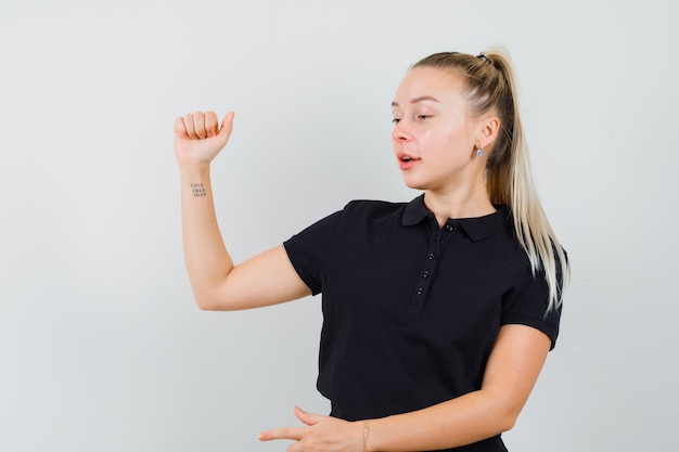 Blonde Dame, die Arm hebt, zeigt zur Seite im schwarzen T-Shirt, Vorderansicht.