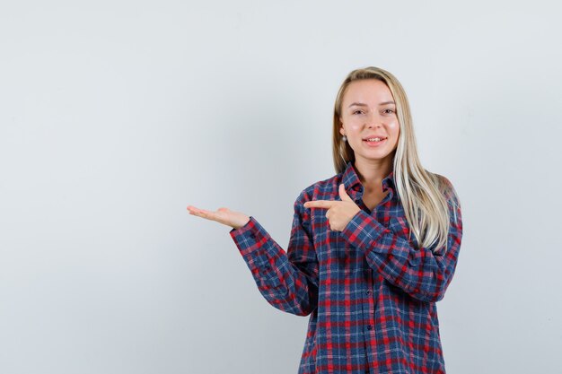 Blonde Dame, die auf ihre Handfläche zeigt, breitete sich im Freizeithemd aus und sah fröhlich aus. Vorderansicht.