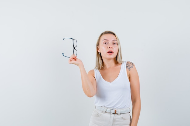 Blonde Dame, die Brille im Unterhemd, in der Hose hält und nachdenklich, Vorderansicht schaut.