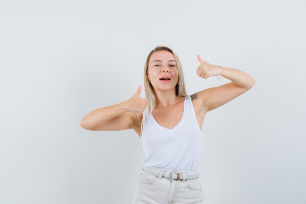 Blonde Dame, die doppelte Daumen in Unterhemd, Hose zeigt und selbstbewusst aussieht. Vorderansicht.