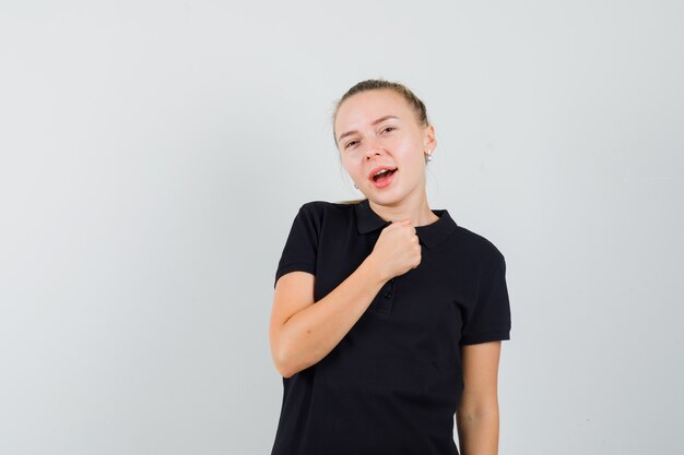 Blonde Dame, die geballte Faust auf Brust in schwarzem T-Shirt hält und glücklich schaut. Vorderansicht.