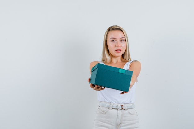 Blonde Dame, die Geschenkbox im Unterhemd, in der Hose präsentiert und niedlich schaut. Vorderansicht.