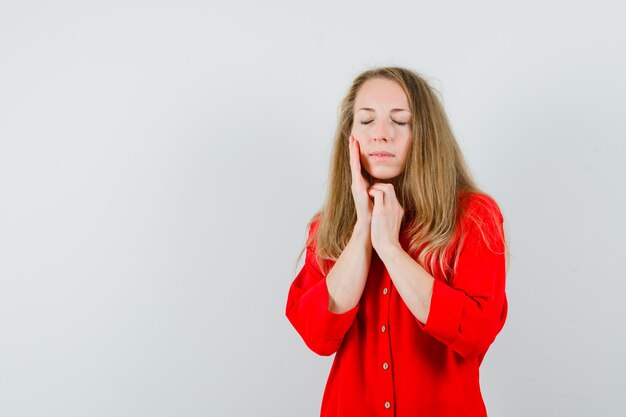 Blonde Dame, die Gesichtshaut auf Wange im roten Hemd berührt und ruhig schaut.